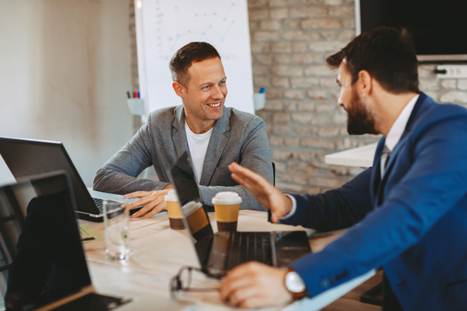 Two businessmen having a friendly discussion at a modern office table with laptops and coffee.