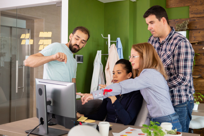 Four colleagues collaborating around a desktop computer in a casual office setting, actively discussing something on the screen.