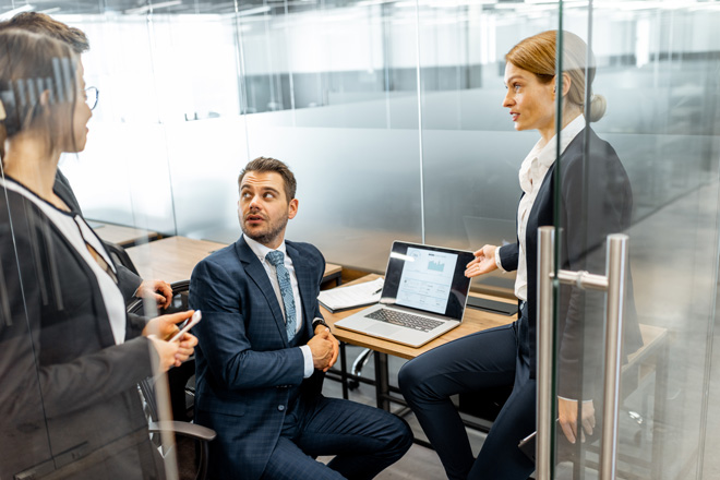 Four business professionals having a discussion in a glass-walled office, with a laptop displaying charts on the desk.