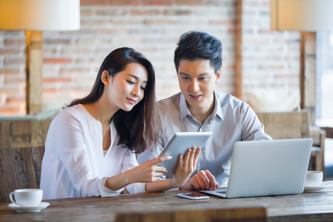Two young professionals sitting at a cafe table, looking at a tablet together with a laptop and coffee cups in front of them.
