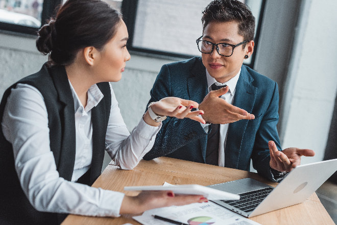 Two business professionals engaged in a discussion at a desk with a laptop and financial documents.